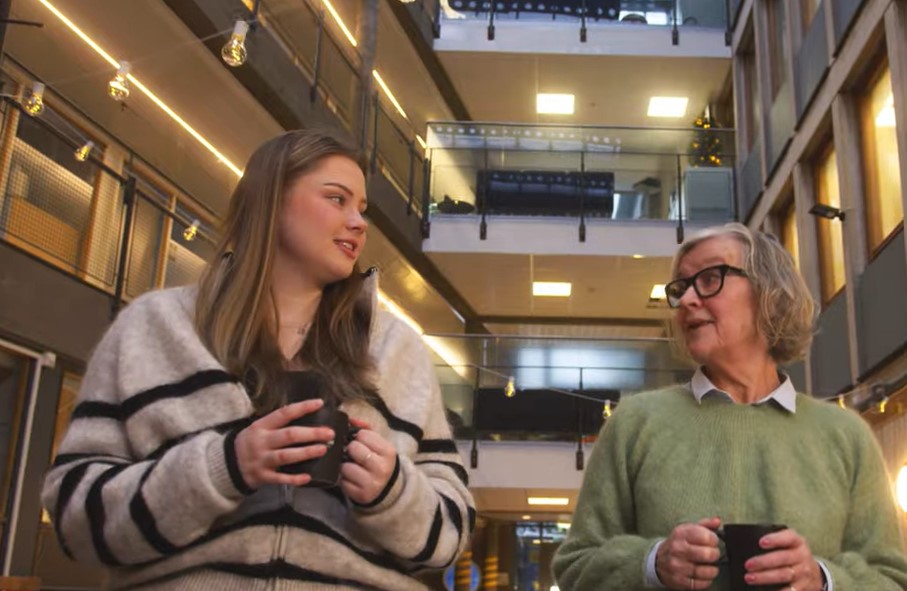 Lærling og mentor med hver sin kaffekopp går i kontorlandskap.