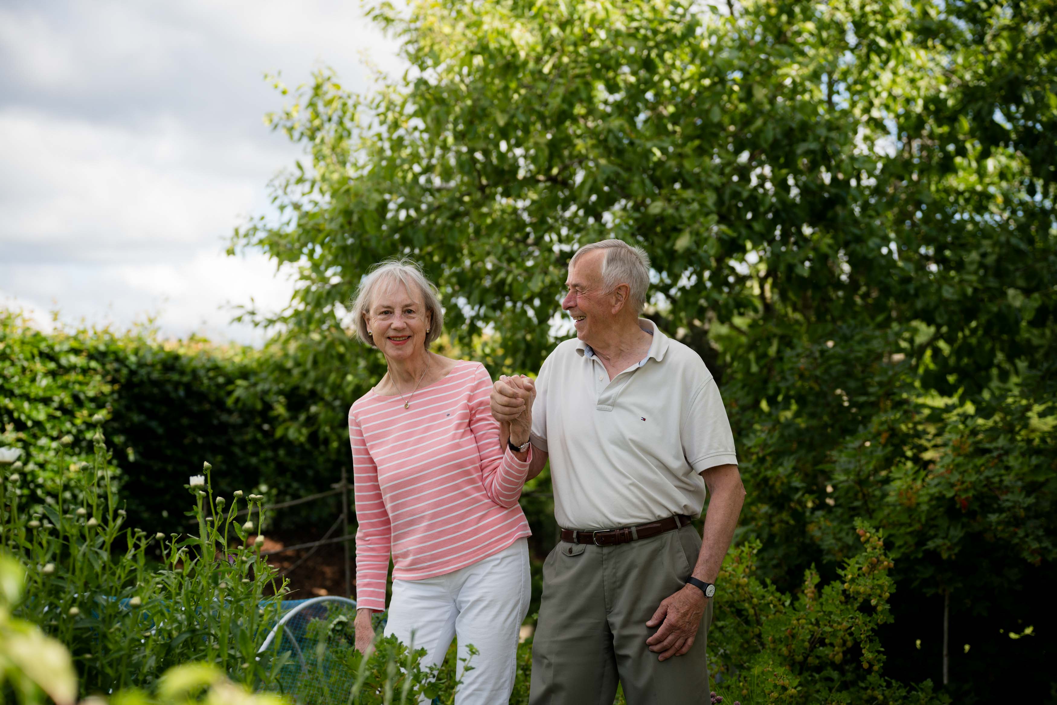 Eldre dame og mann som holder hender og smiler i en grønn og frodig hage.