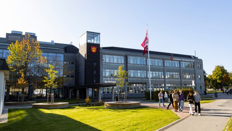 Inngangspartiet til skolen, elever går ut av bygget, det er grønne blader på trærne.