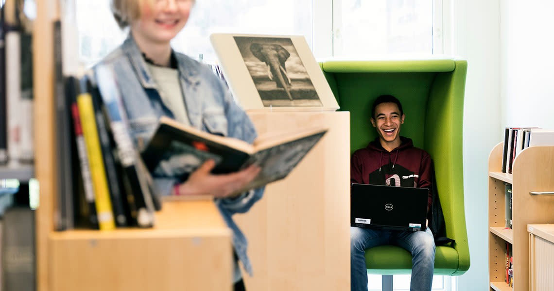 En smilende elev leser i bok og en elev i stol som ler