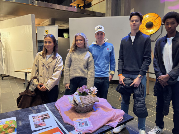 To jenter og tre gutter står bak et bord med brosjyrer og en blomst.