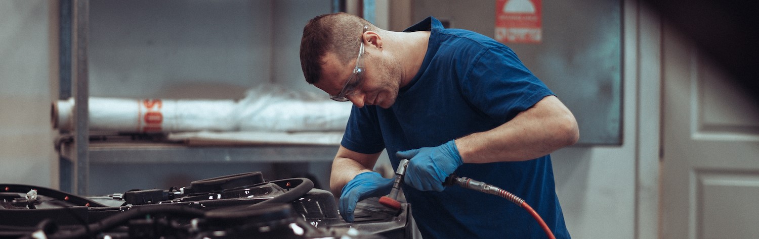 Mann med t-skjorte, vernebriller og gummihansker arbeider med er trykkluftdrevet slipeverktøy på en stor maskin.