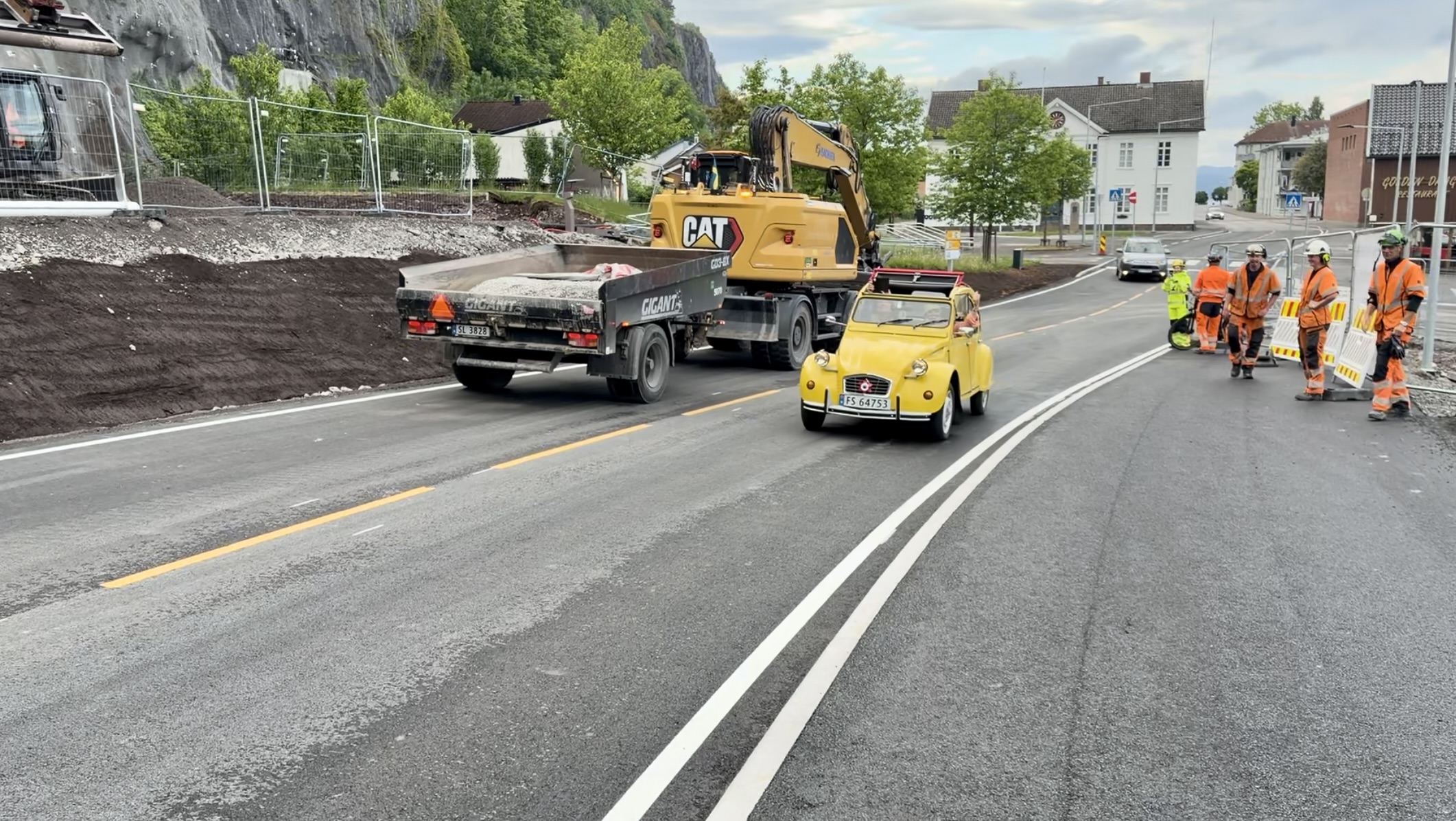 Den første bilen som kjører på den nye veien eer en gul Citroen 2CV. Gravemaskin og anleggsarbeidere står på fortauet og ser debn første bilen som kjører.