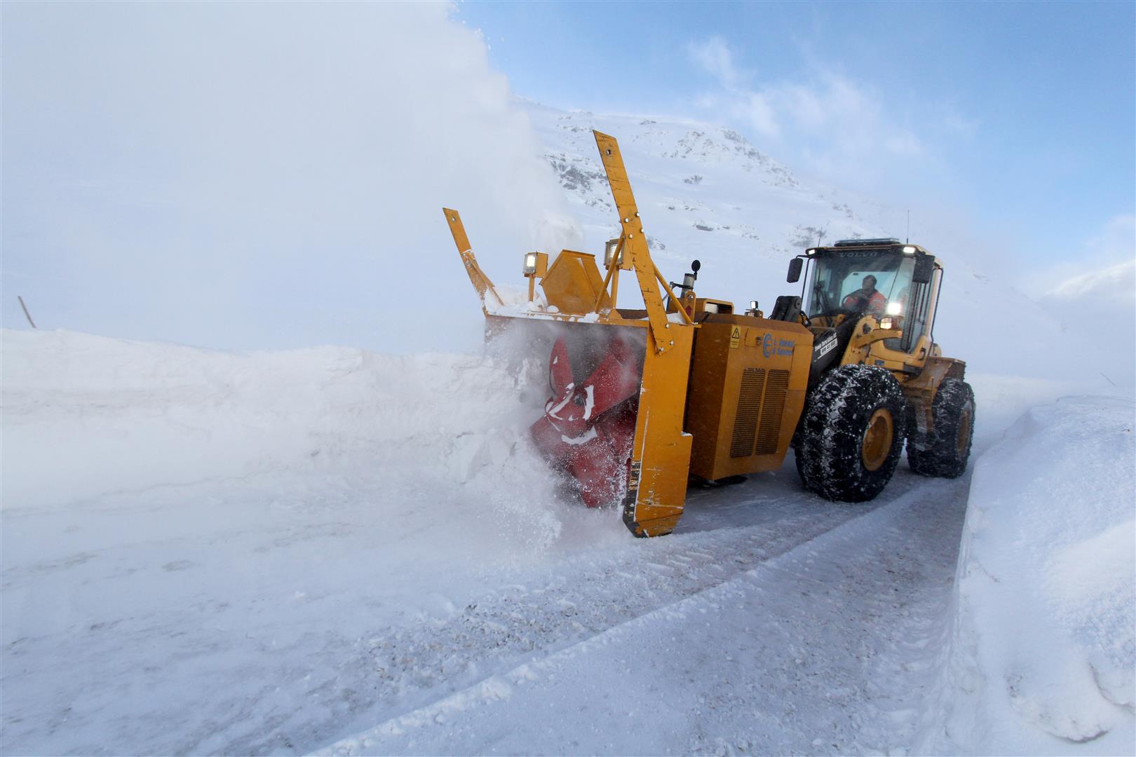 Snøfreser kjører mot synsvinkelen til fotografen. Snøføyken står rundt maskinen. Man skimter fjell i bakgrunnen.