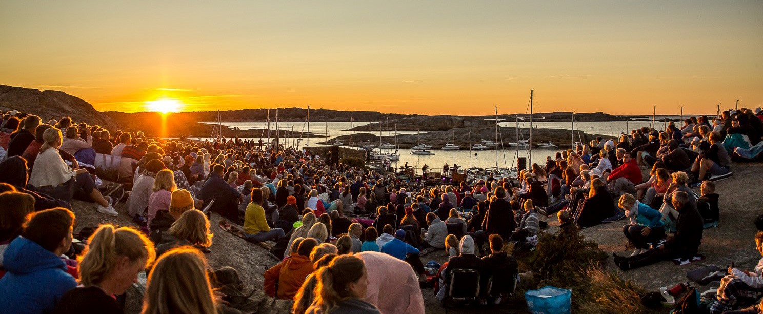 Utendørs sommerkonsert. Mye publikum, og sol og hav i bakgrunnen.