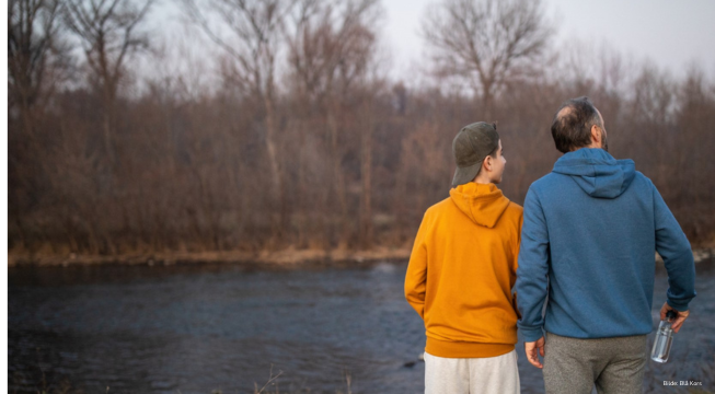 Far og barn ved vannet, med ryggen til kamera.