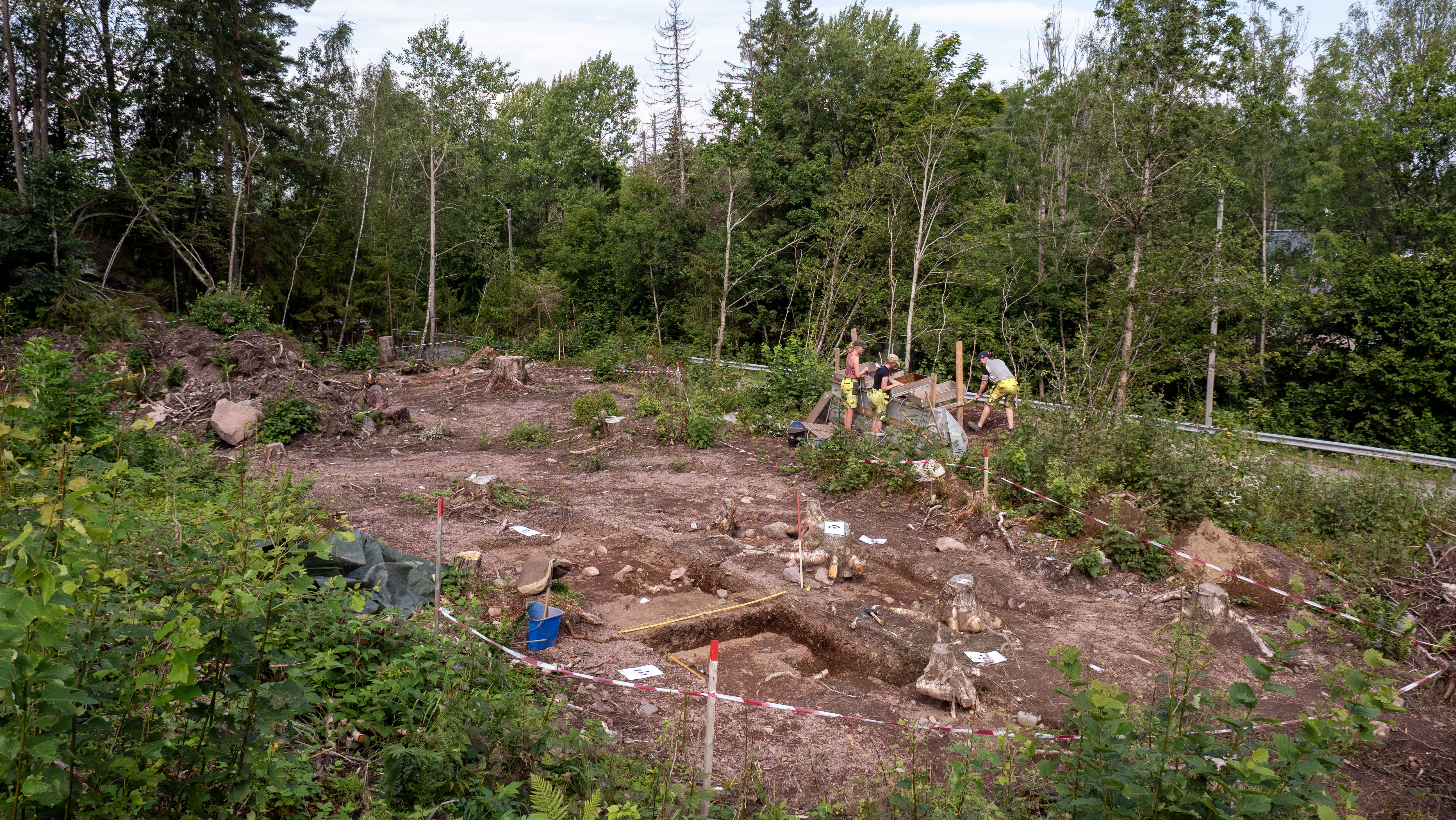 Bildet viser en oversikt over stedet det er gjort arkeologiske utgravninger sommeren 2025. I bakgrunnen i bildet ser vi tre arkeologer som står og jobber.