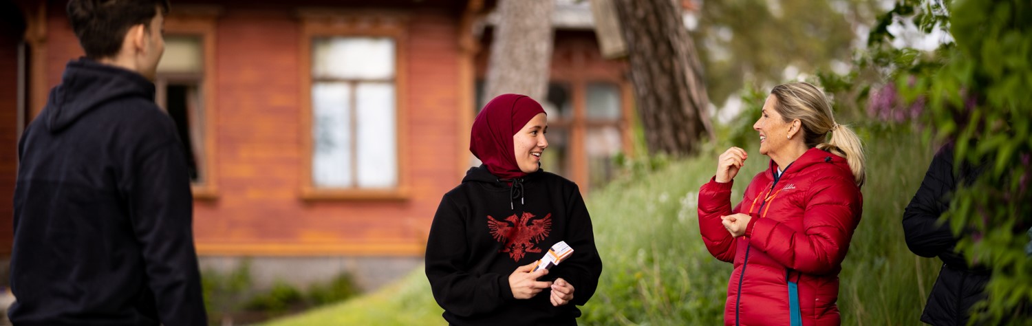 To kvinner snakker og smiler utendørs. En ung mann ser på.