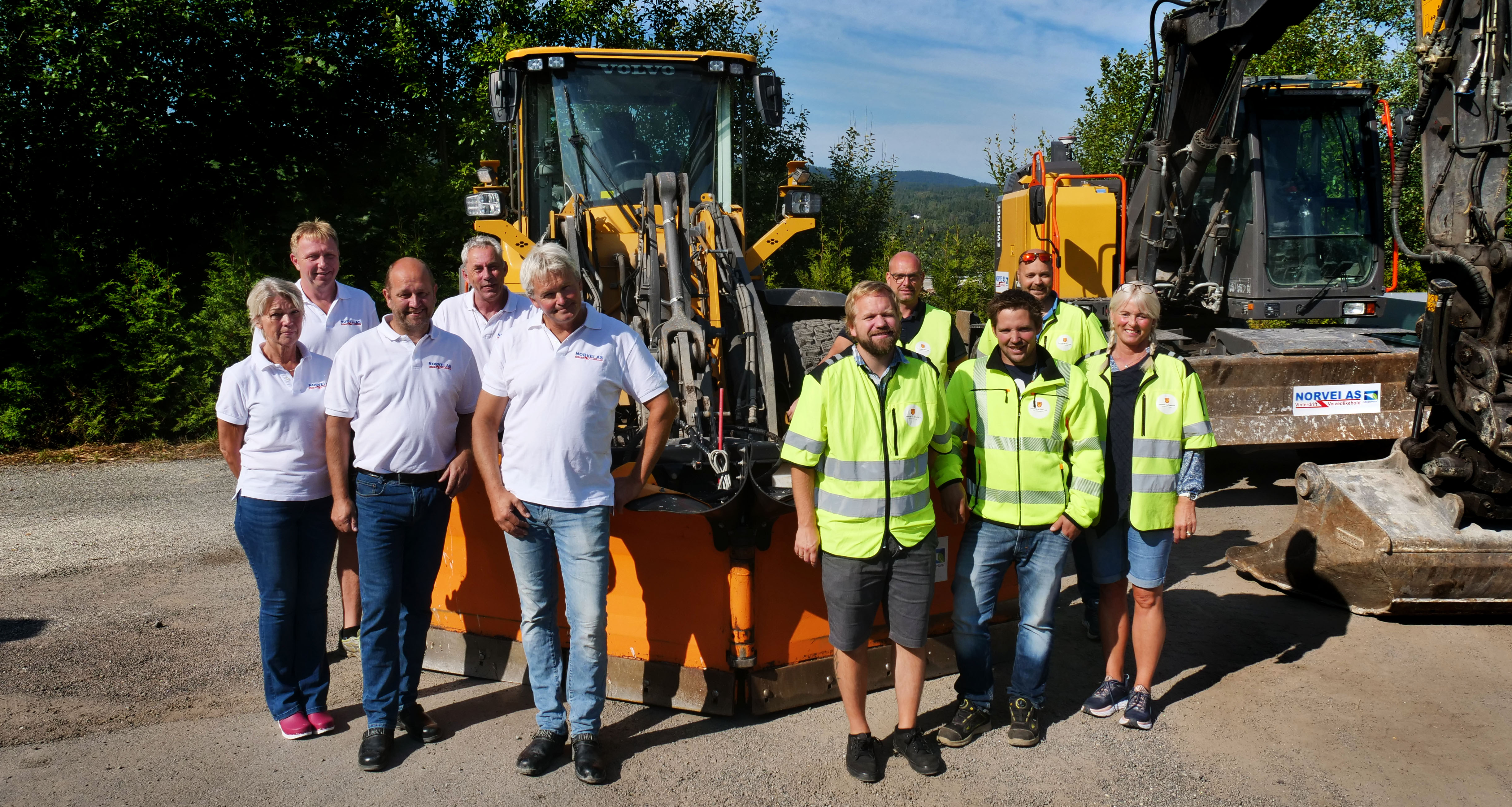Gruppe mennesker fra Norvei AS og fylkeskommunen sett forfra, stående foran brøytebil og gravemaskin.
