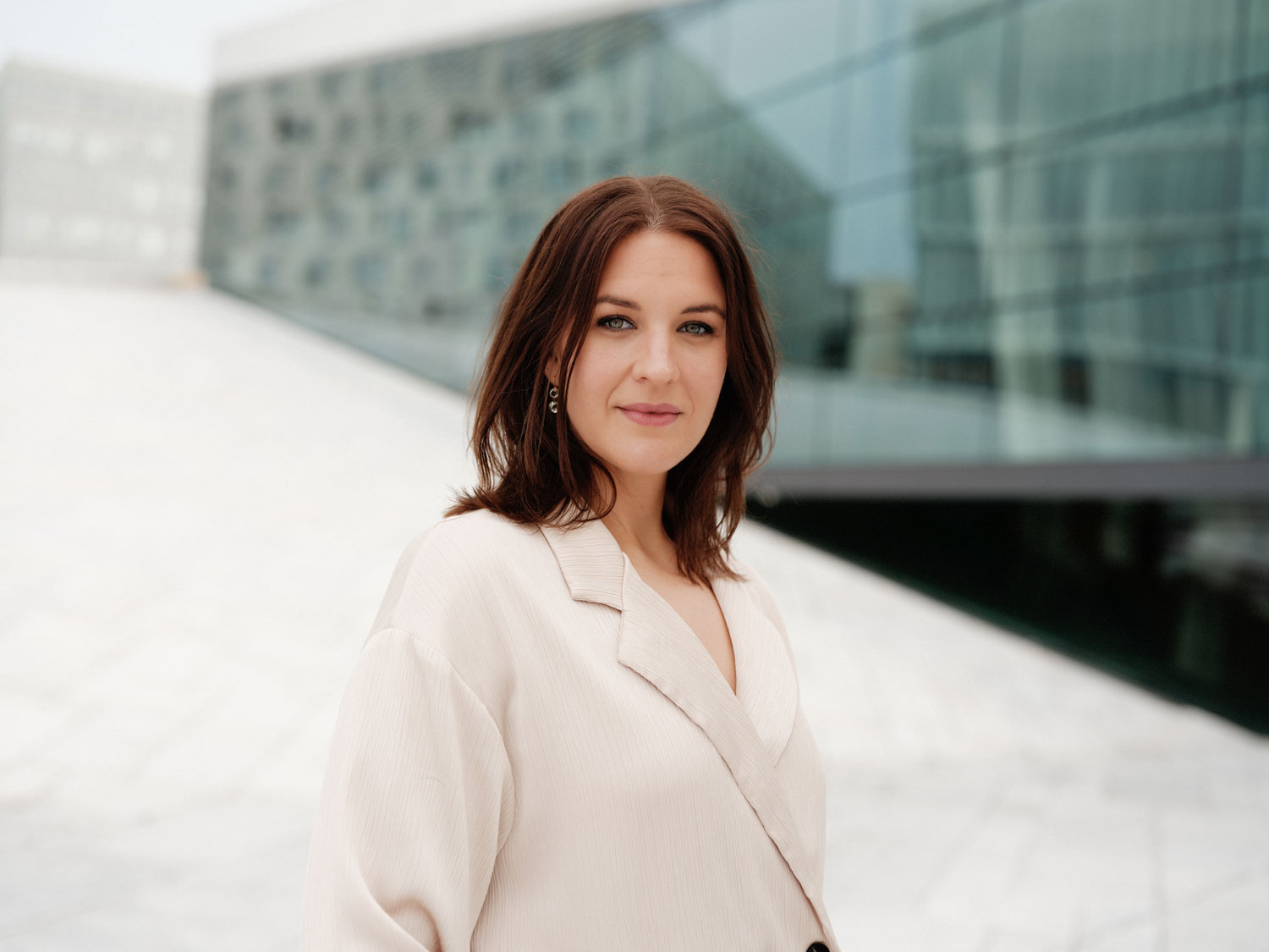Lise Davidsen iført en hvit dressjakke. I bakgrunnen: Taket på operaen i hvit marmor og vinduer som speiler Deichmanske hovedbibliotek og andre hvite bygg.