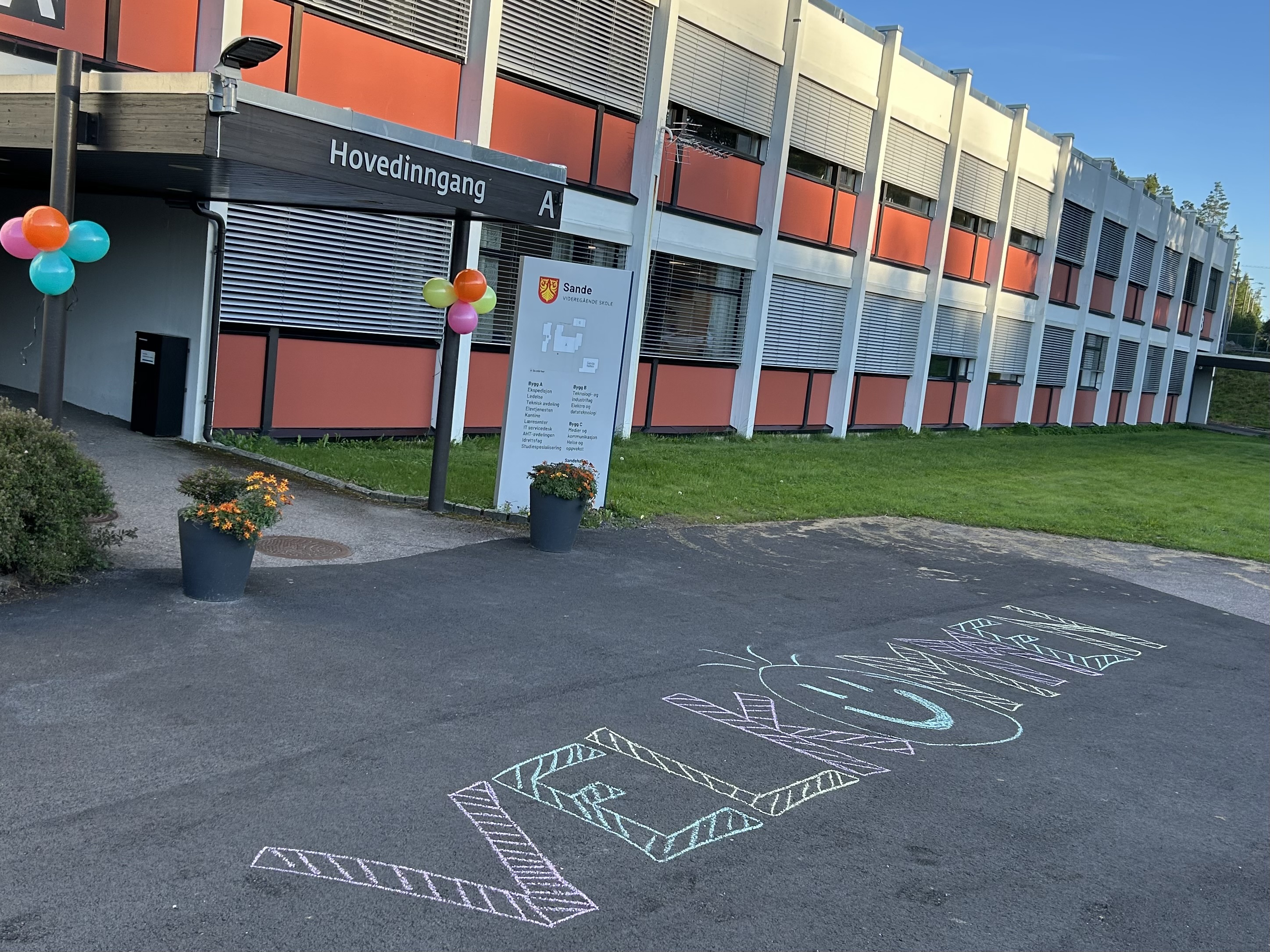 Skolebygning med "velkommen" skrevet med kritt på asfalten foran hovedinngangen
