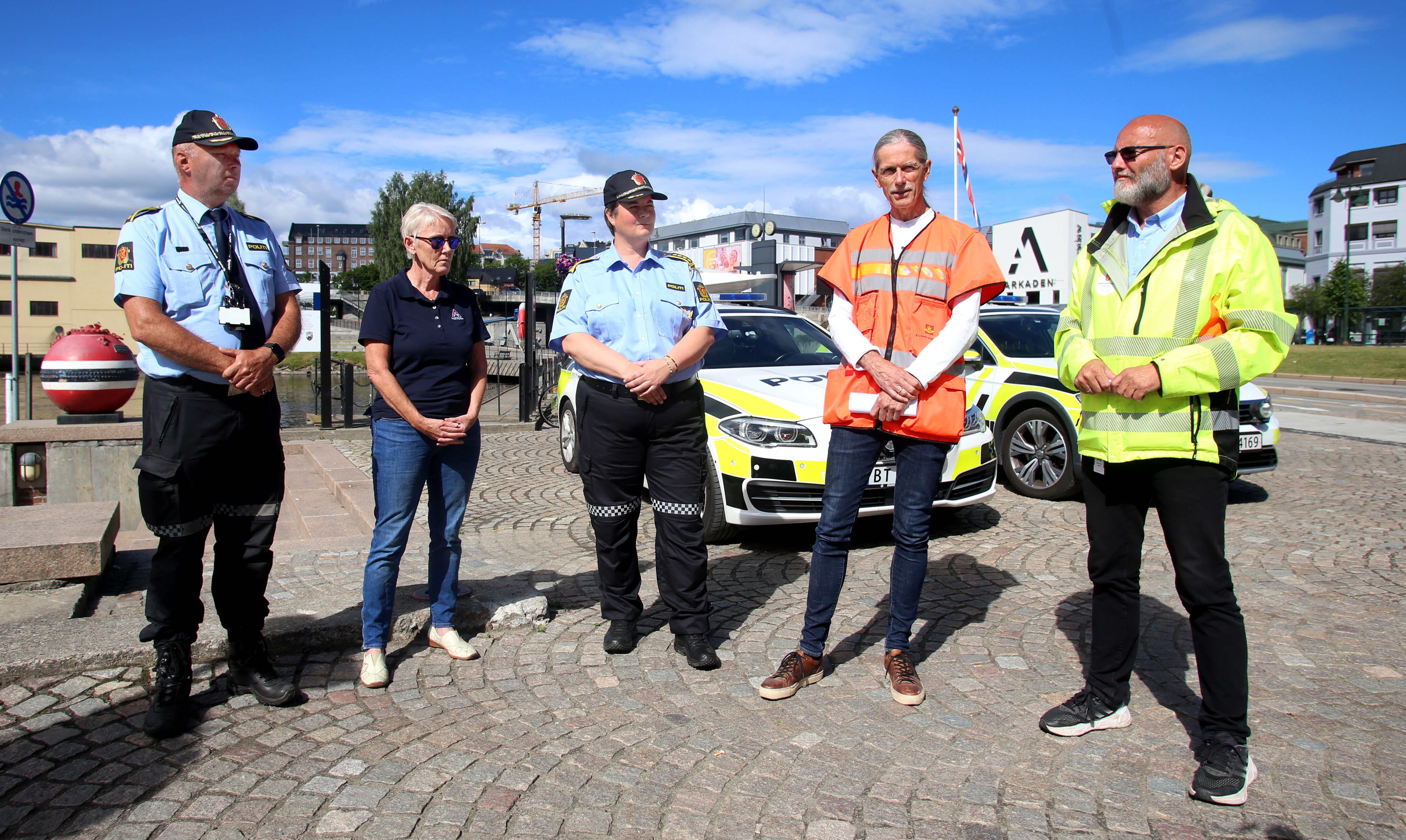 Fem personer oppstilt på rekke, med politibiler i bakgrunnen. To av personene har politiuniform. De andre har også tøy som representerer hvor de kommer fra.