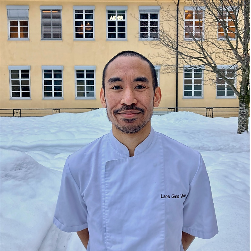 Mann i kokkejakke står ute i snøen foran en gul bygning og ser i kamera.